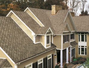 Large home with beige house siding, two white pillars on porch, and brown asphalt shingle roofing. 