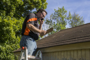 Contractor on ladder with cell phone calculating hail damage repair costs for homeowner.